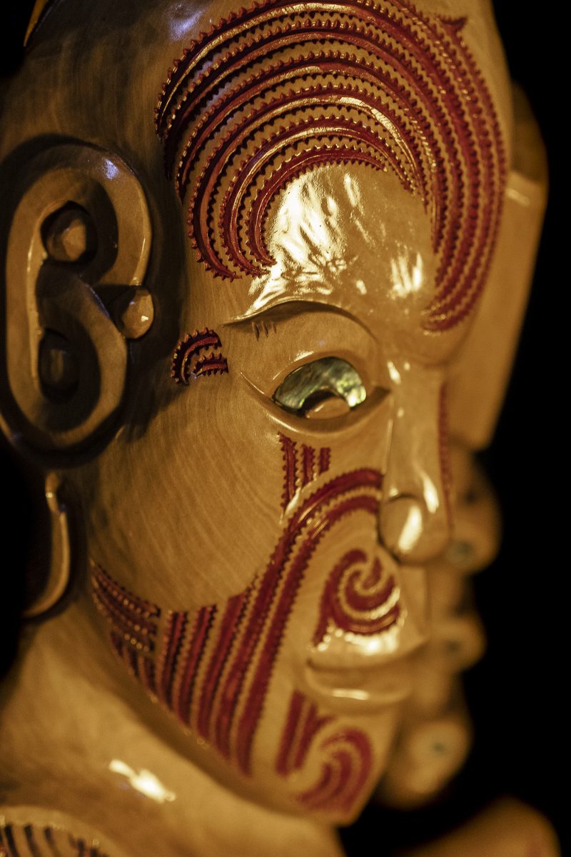 Whakairo by Kade Cowper: a carved face in profile with a pāua eye, its moko cut deep and picked out in red.